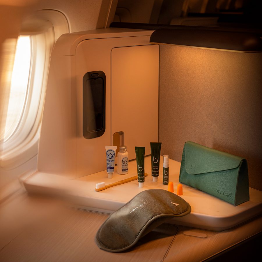 A lifestyle shot of the new Cathay Pacific Bamford amenity kit on a shelf next to an aircraft window, featuring a stylish case, eye mask, toothbrush, ear plugs and toiletries in recyclable packaging