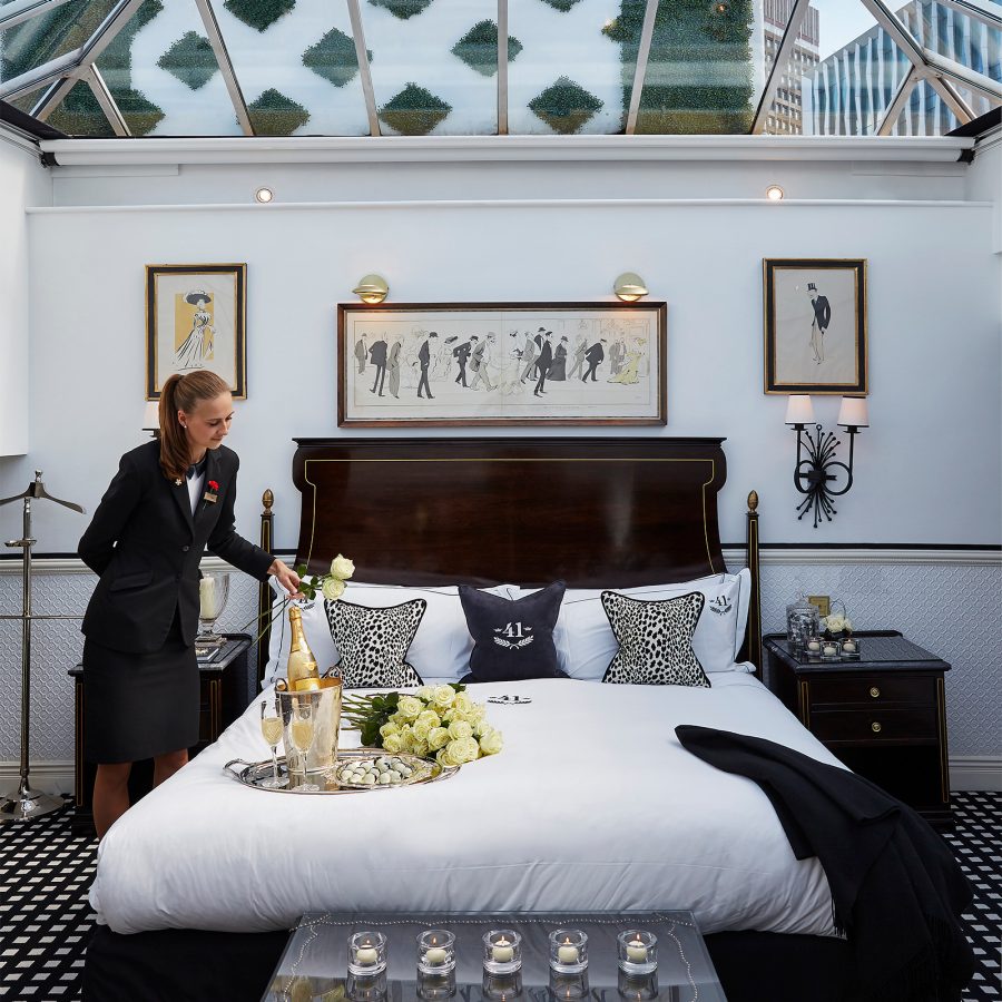 An inside view of a luxe bedroom suite at Hotel 41 with a skylight view and staff decorating the bed with flowers and champagne.