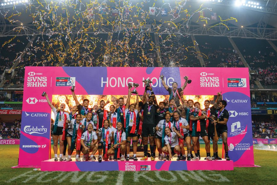 A group of rugby players celebrating and cheering with trophies on the field in Hong Kong infront of a banner and streamers in the sky.
