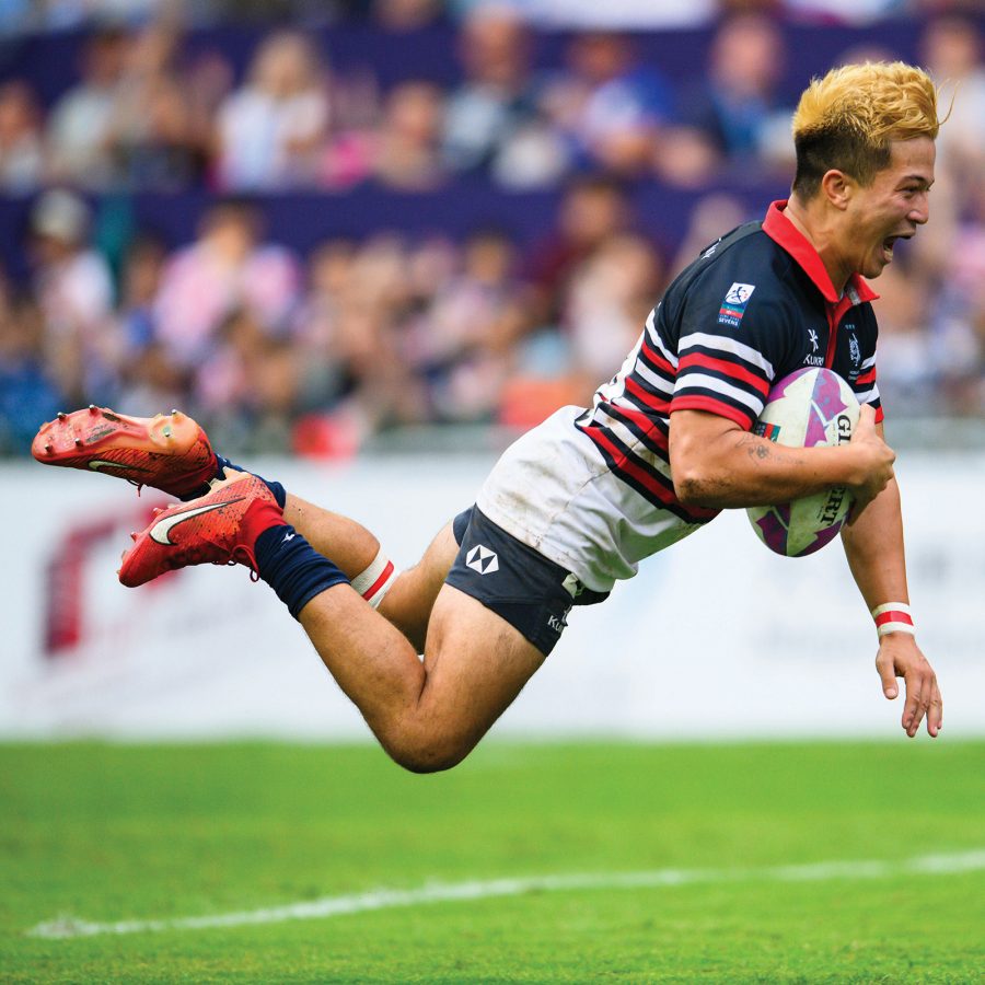 A rugby player holding a rugby ball and leaping forward on a field.