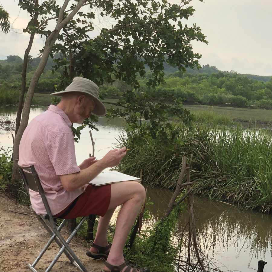 A man sitting on a camping chair next to a river sketching nature at Elsie Evans Art Retreat in Ryong, Thailand