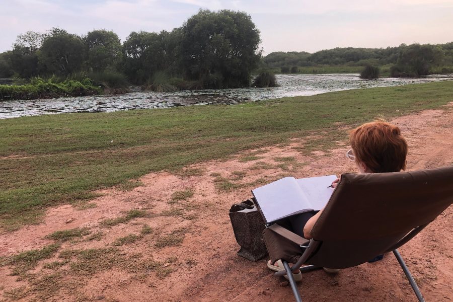 A woman sitting on a camping chair by the river, looking out and sketching nature at Elsi Evans Art Retreat in Ryong, Thailand