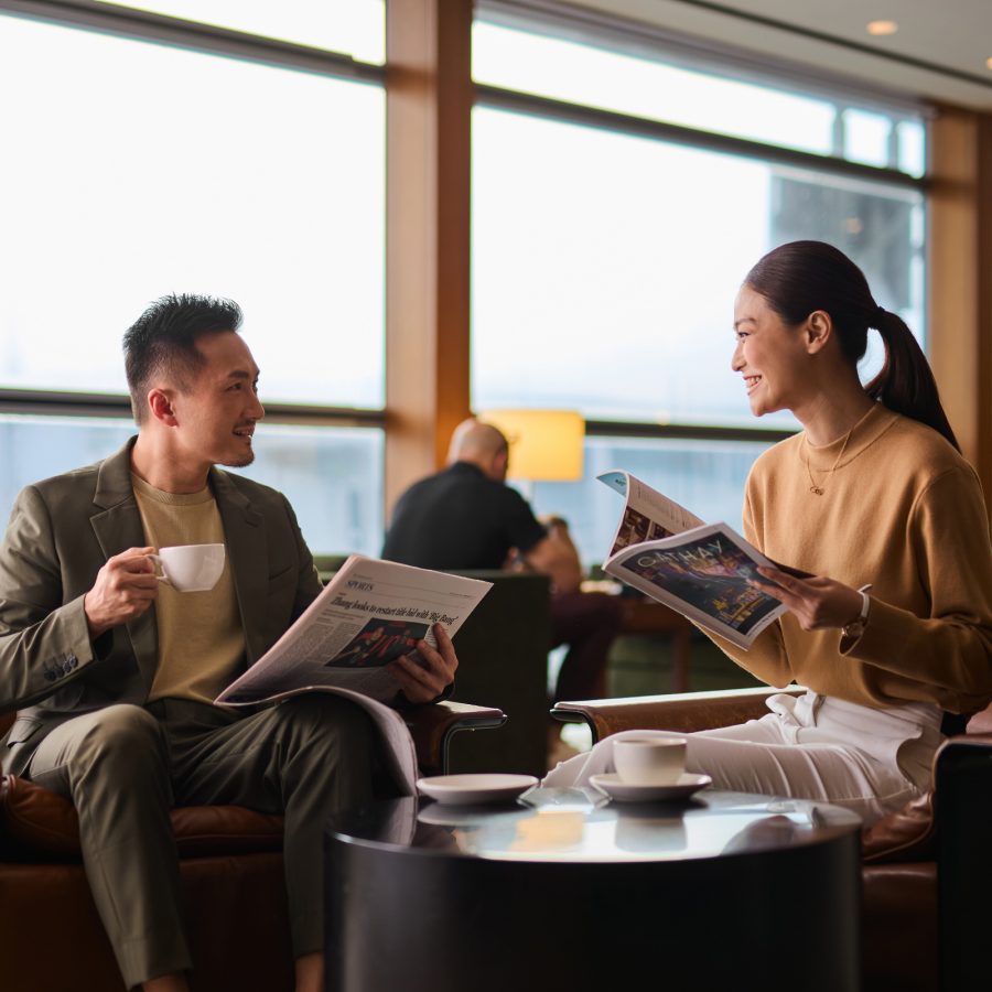 A man holding a cup and newspaper smiling at a woman holding a Cathay magazine a Cathay Pacific lounge.