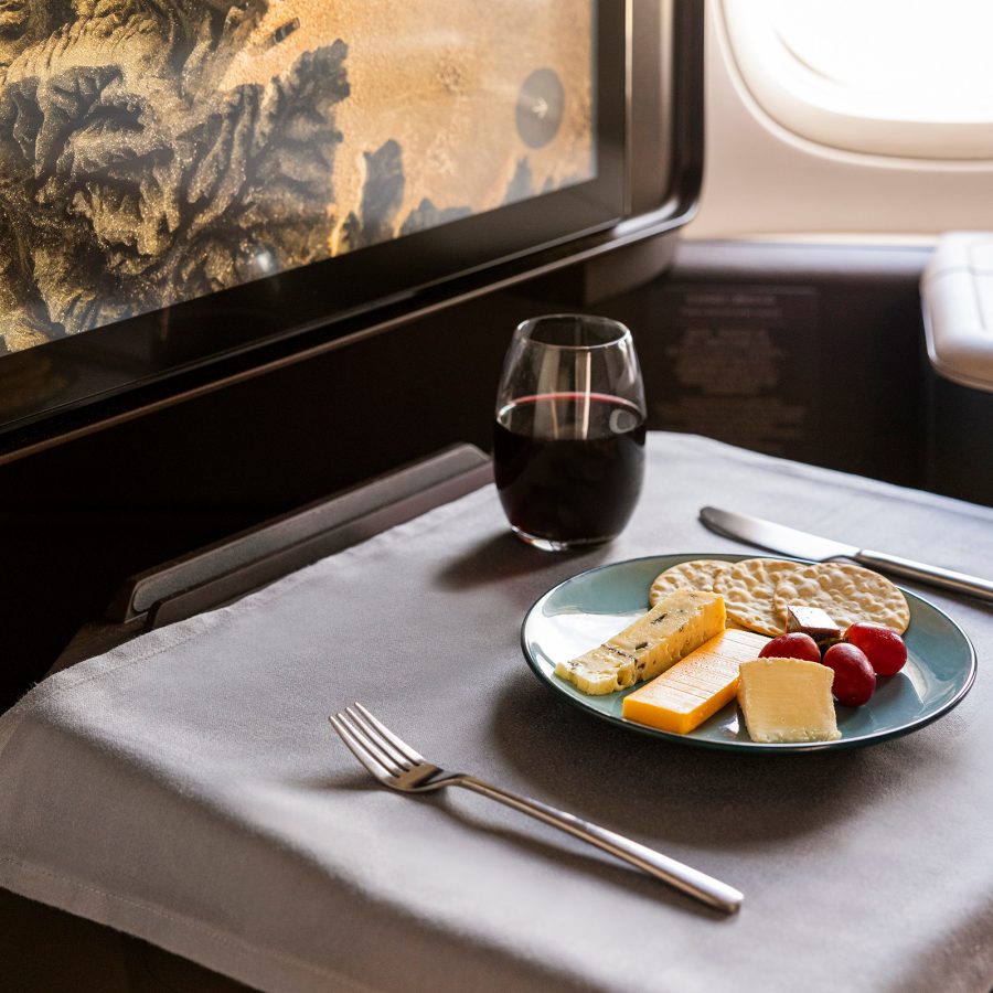 At a First class passenger seat on a Cathay Pacific aircraft, a fold-out tray is laid with a tablecloth, a plate of various cheeses, crackers and fruit, and a glass of red wine.