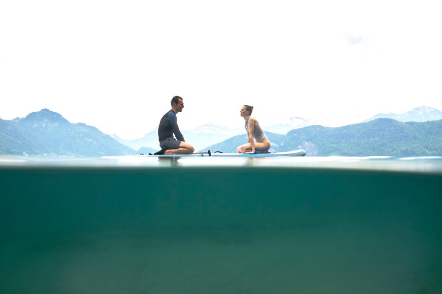 A couple of man and woman on surfboards outdoors facing each other with a view of the mountain range in the backdrop during daytime.