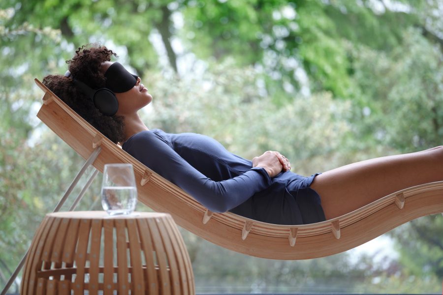 A woman taking a nap on a wooden daybed while wearing an eye mask with green trees in the backdrop during daytime.