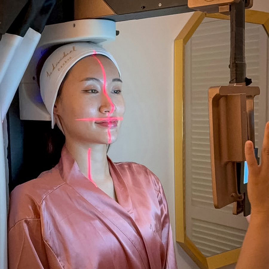 A woman in a pink robe sitting in front of a 3D skin camera with laser beams on her face. 