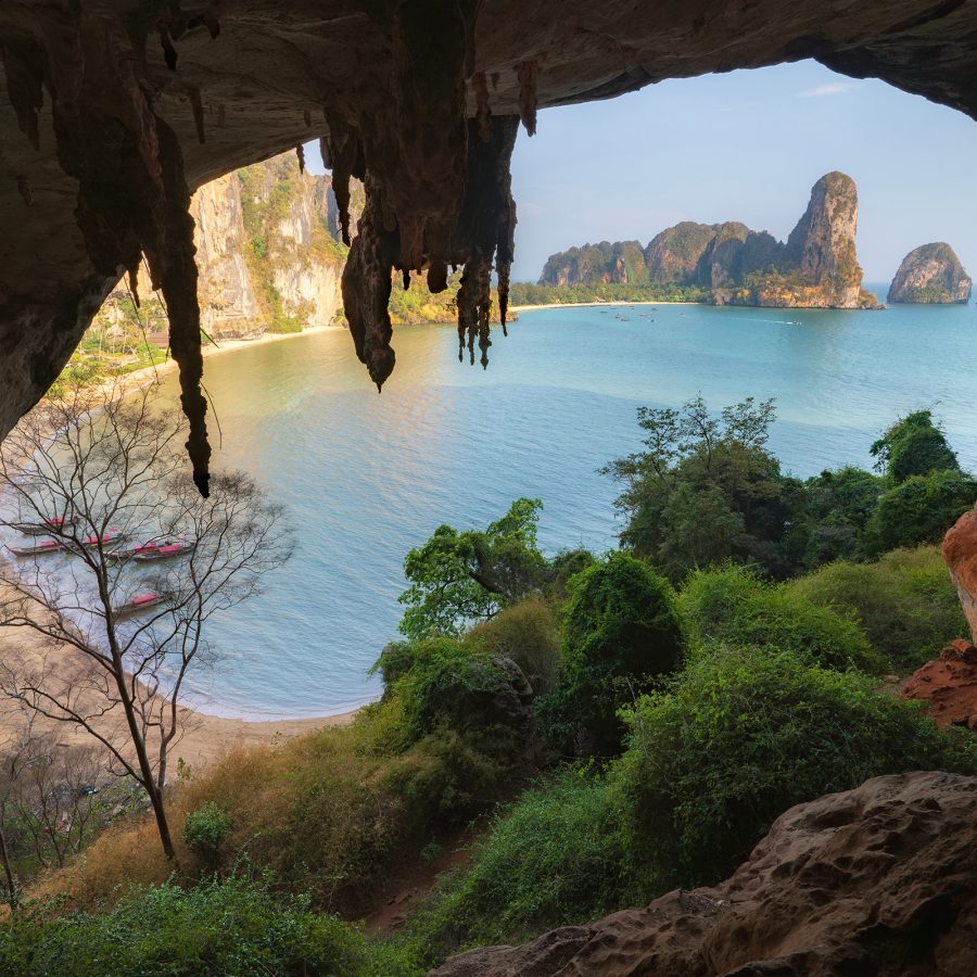A view of Railay Beach in Krabi, Thailand, seen from inside a cave with lush green foliage on a sunny day.