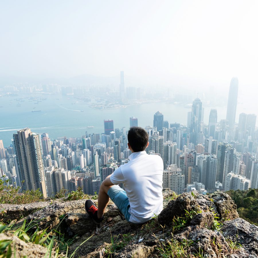 A man sitting at the top of a hill with a view of the city’s skyscrapers and harbour in Hong Kong on a sunny day.