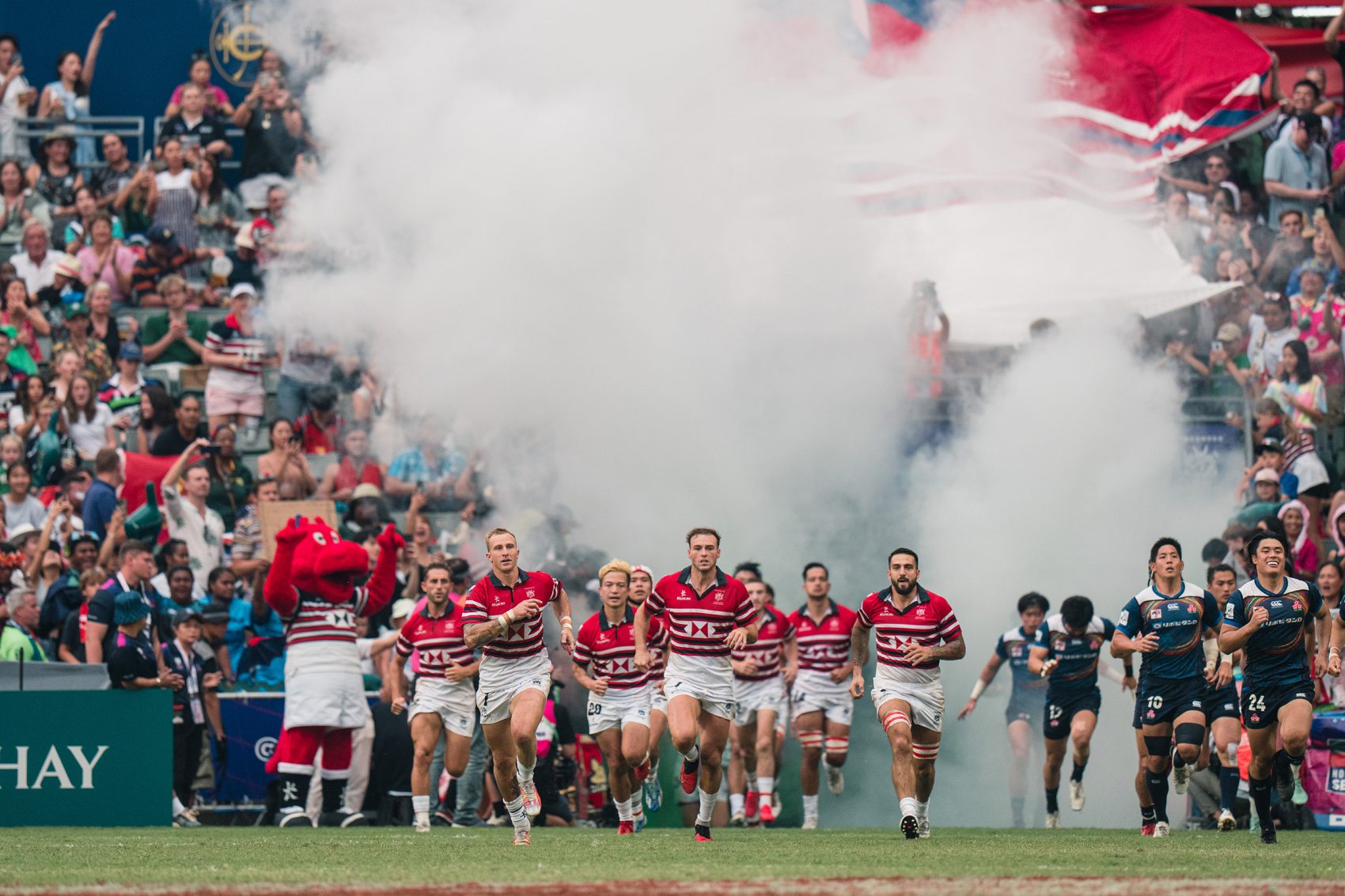 Rugby players run onto the pitch against a backdrop of fog and crowds.