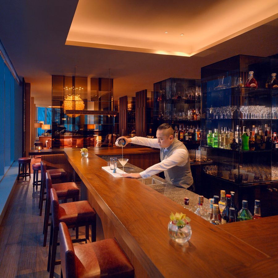 Bartender pours a cocktail at The Roof Bar with a view of the skyline through glass windows.