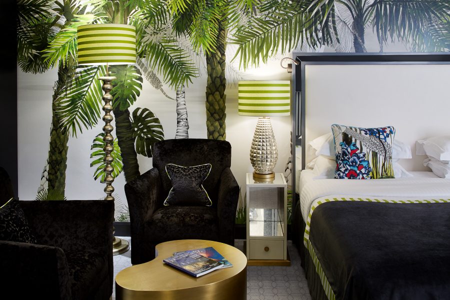 The interior of a hotel room shows a bed with a colourful pillow, palm tree-themed wallpaper and playful furniture.