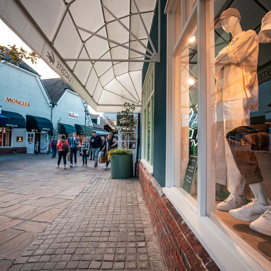A glass storefront features two mannequins sporting various types of apparel as pedestrians pass by at a Bicester Village outlet.