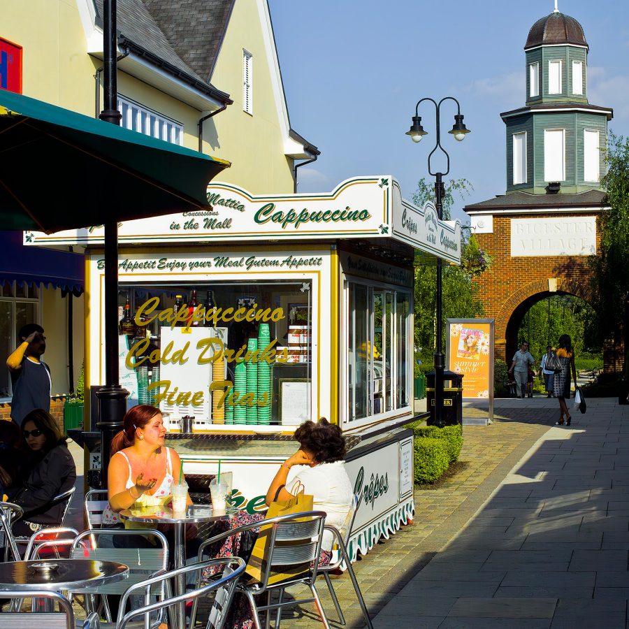 Two women eat and chat at a table outside a coffee stand at Bicester Village. 