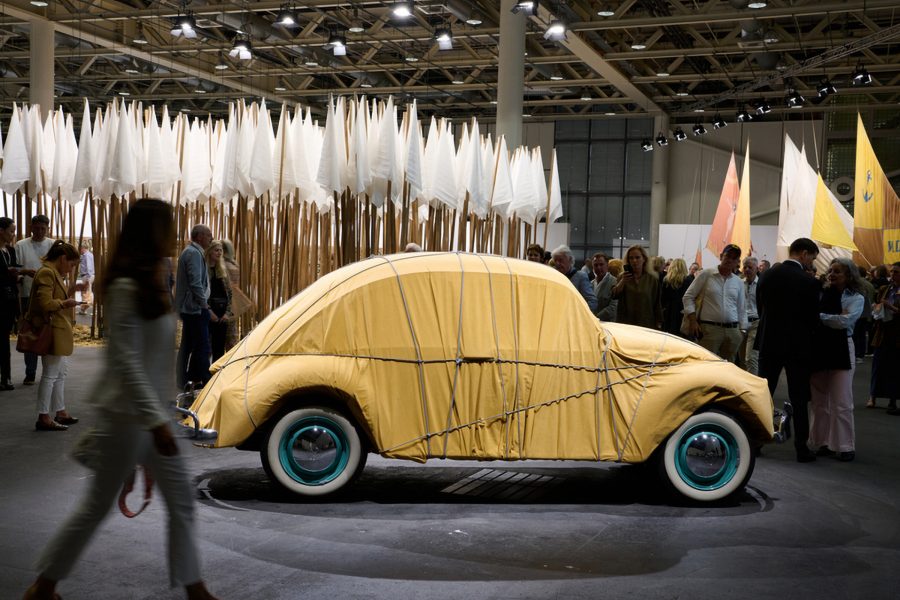 A car covered in a yellow sheet in front of white flags as people look on at Art Basel in Switzerland.