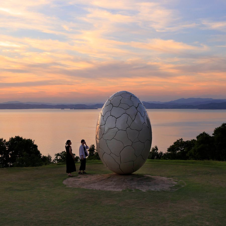 A large egg sculpture on a patch of grass in front of the water at sunset.