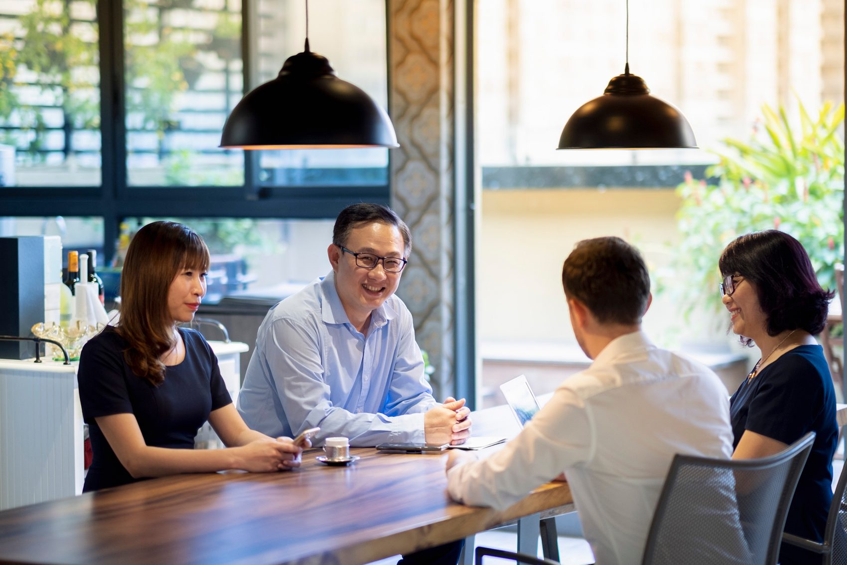 A group of people chat at a wooden desk under two hanging pendant lights.