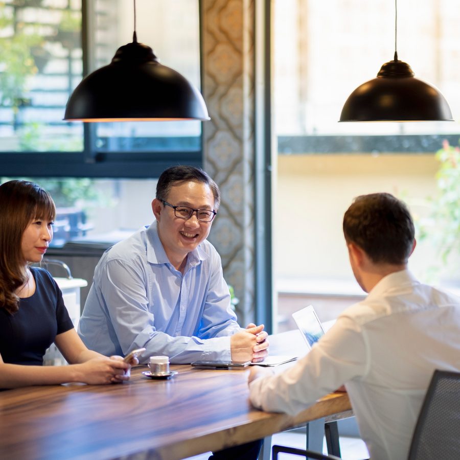 A group of people chat at a wooden desk under two hanging pendant lights.