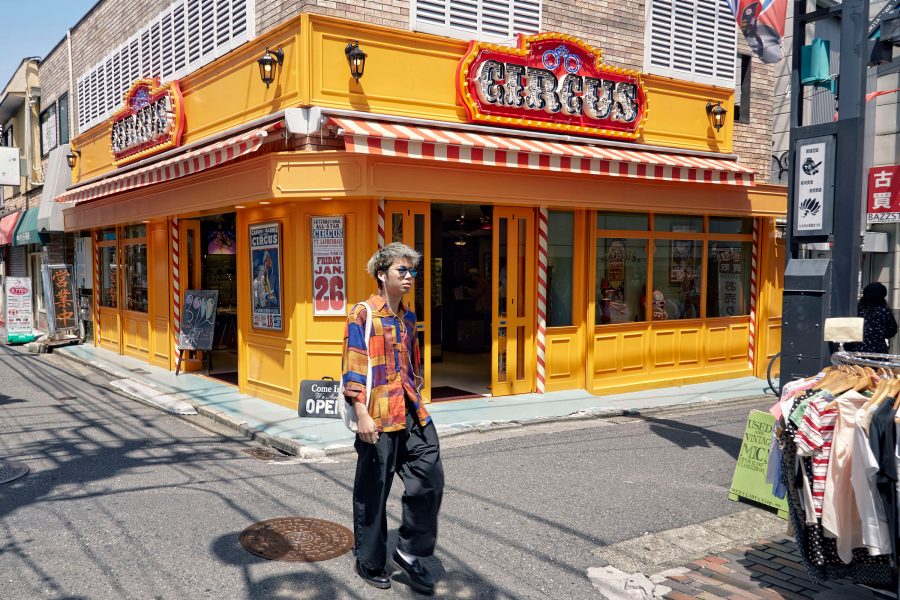 A young man in a colourful shirt walks past the yellow storefront of Circus at Shimokitazawa in Tokyo in the daytime.