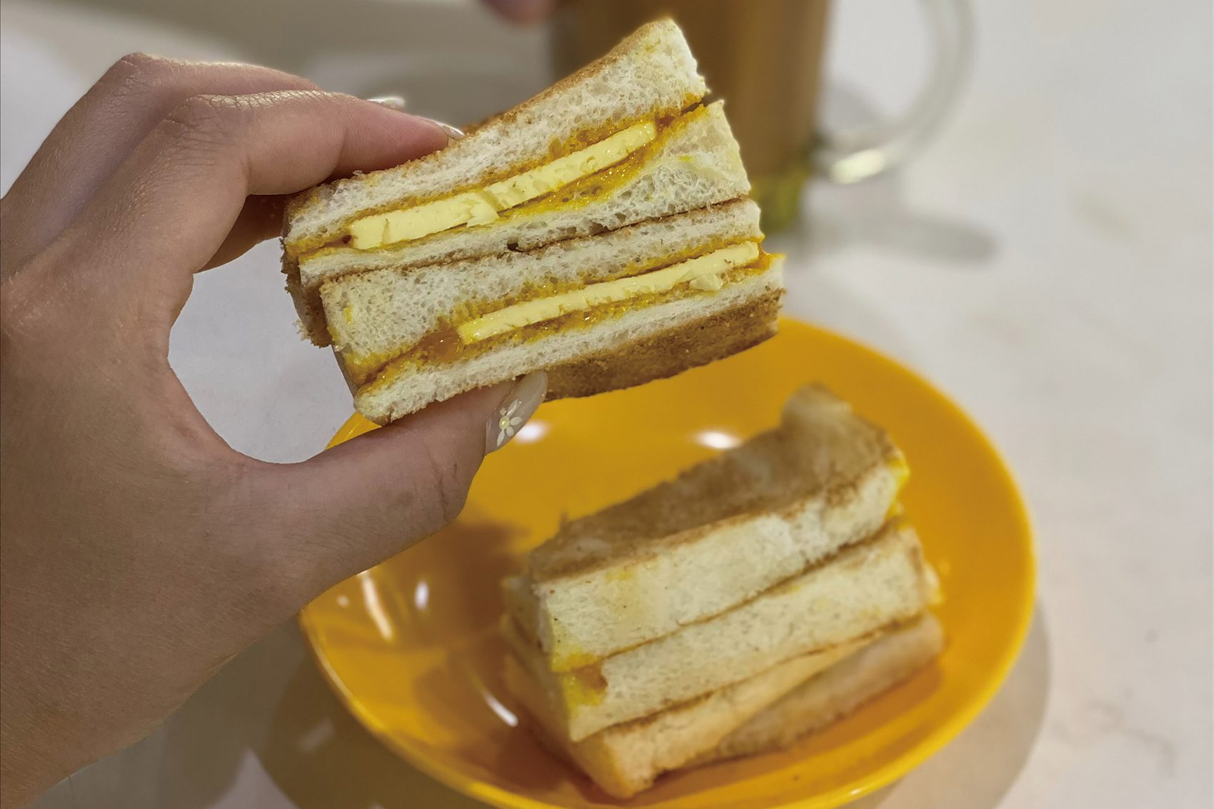 A hand holds a slice of toasted sandwich filled with kaya spread and chunks of butter