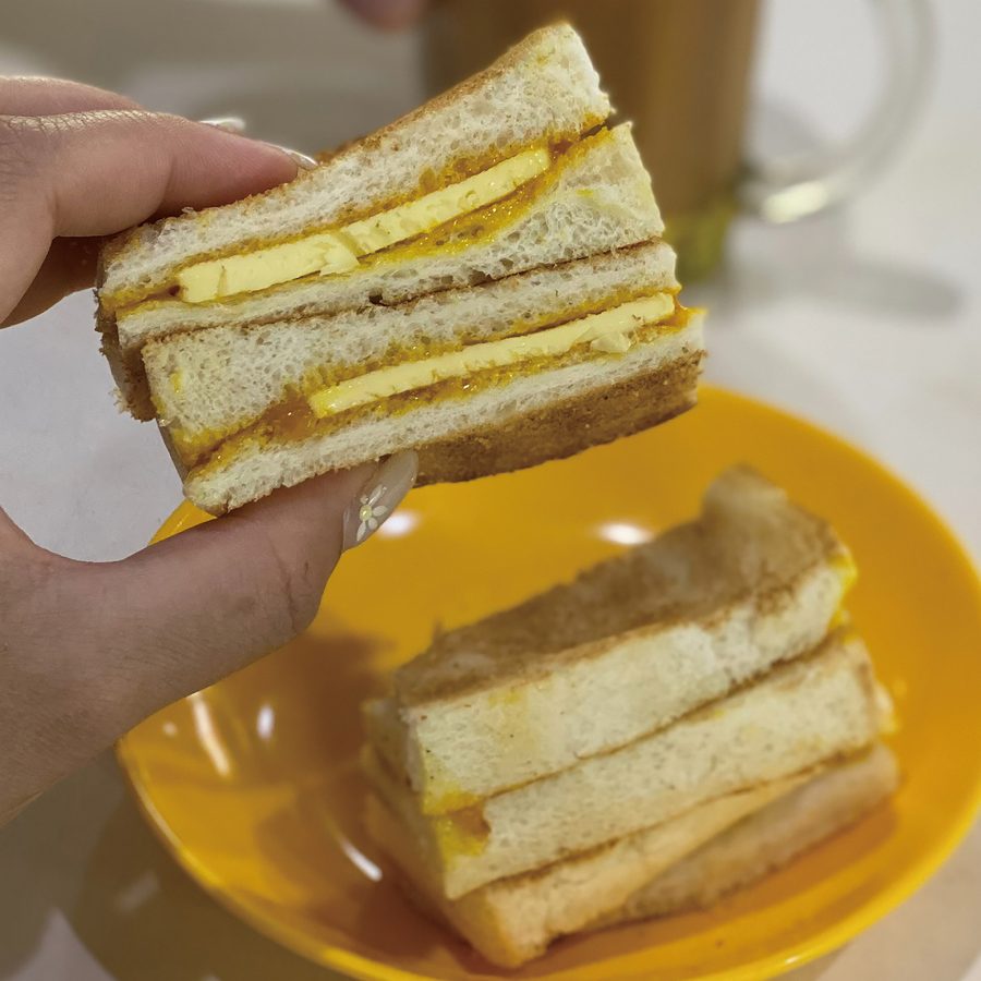 A hand holds a slice of toasted sandwich filled with kaya spread and chunks of butter