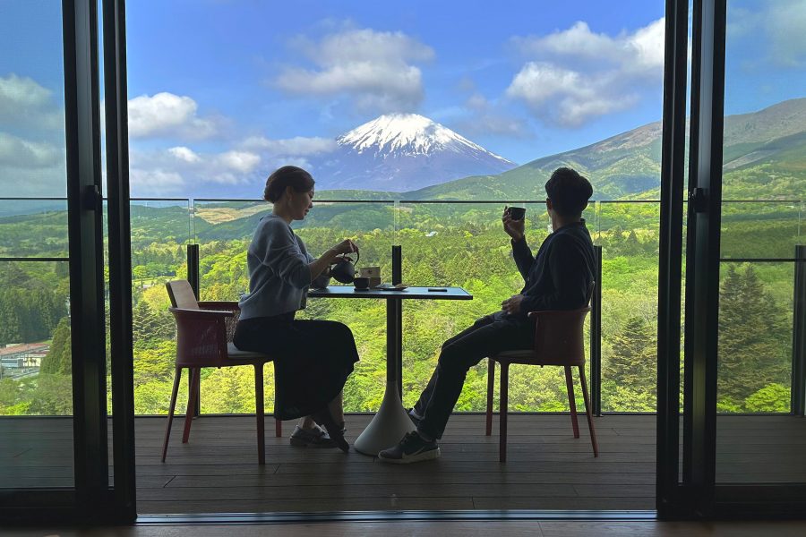 A couple sits on a balcony drinking coffee with a view of Mount Fuji in the background. 