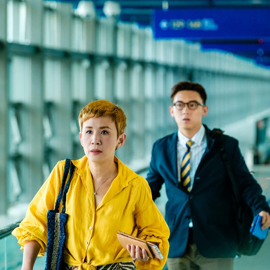 This scene from Love Lies shows middle-aged woman Veronica (played by Sandra Ng) walking along a travelator at Hong Kong International Airport. Behind her walks a young man in a suit, Joe (played by MC Cheung Tin-fu).
