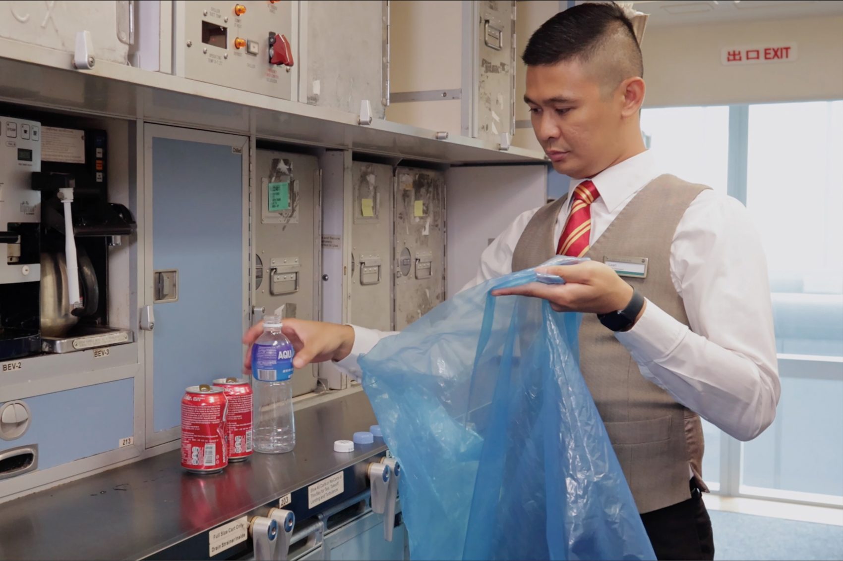 A male Cathay Pacific cabin crew placing an empty plastic bottle into a blue plastic bag, designated for recycling.