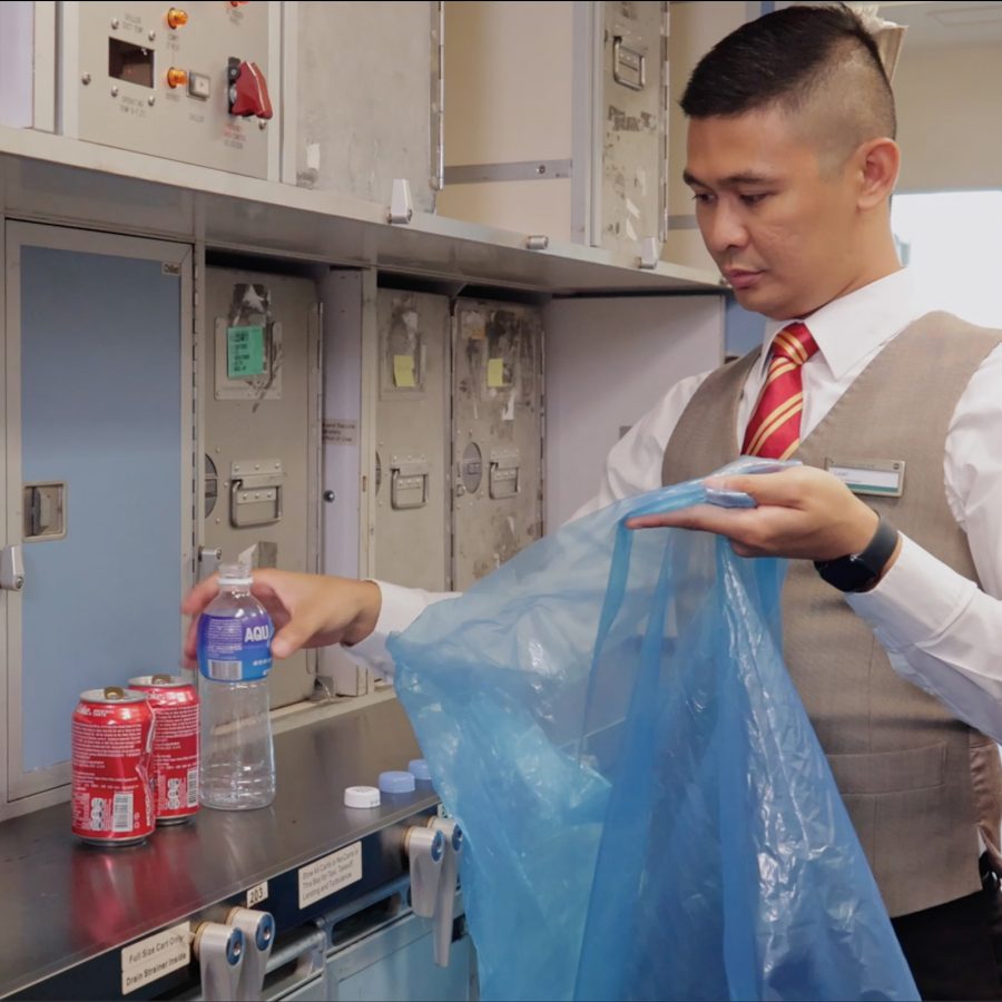 A male Cathay Pacific cabin crew placing an empty plastic bottle into a blue plastic bag, designated for recycling.