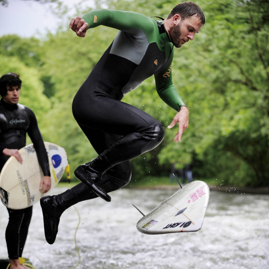 A male surfer leaps into the Eisbach with a surfboard at his feet.