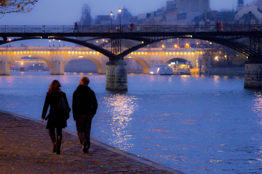 A couple walk along the Seine at night as bridges along their path light up.