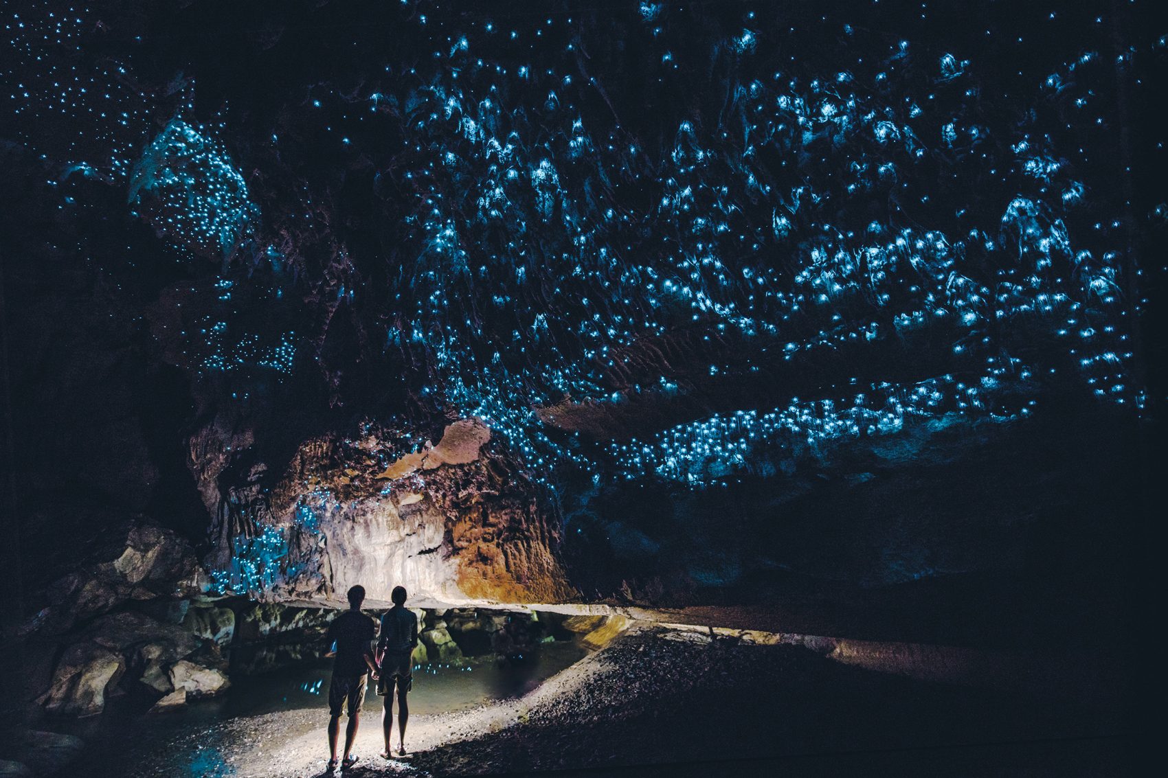 Two people walk through the Waitomo Glow Caves and the glowing blue worms across the ceiling.