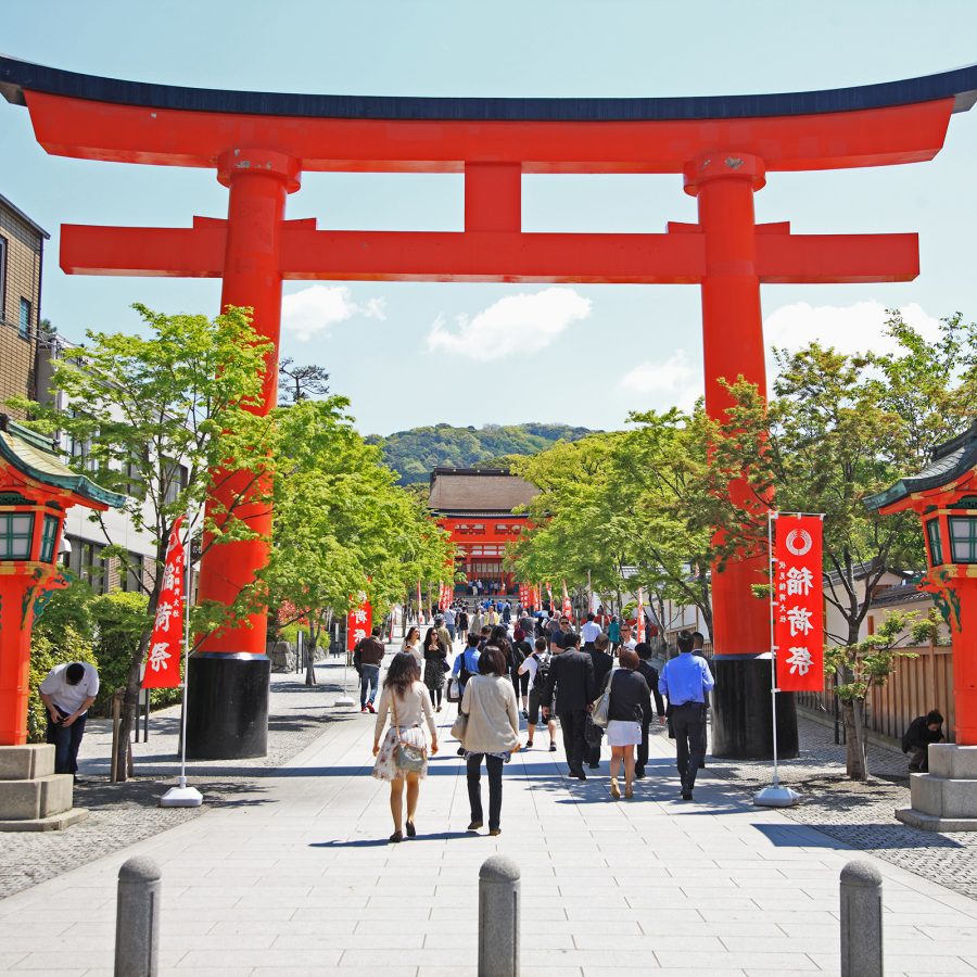 Crowds of travellers going through the entrance of  Fushimi Inari Taisha temple grounds in Kyoto on a sunny day.