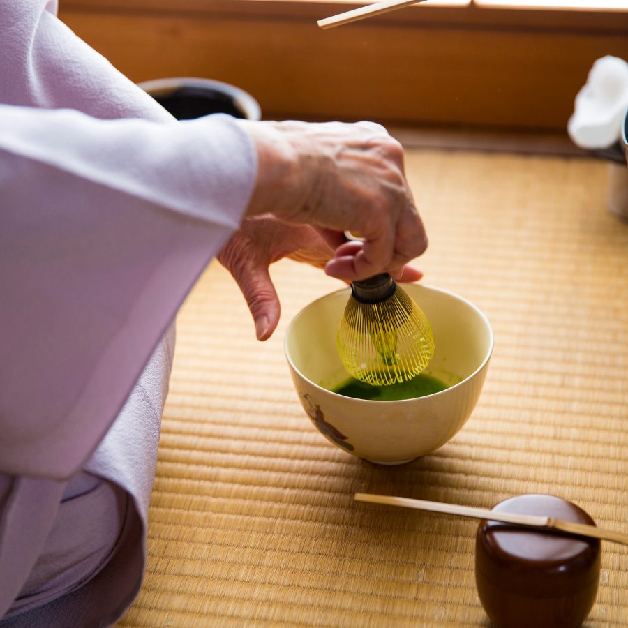 A closeup of a person wearing a hakama kneeling on a bamboo mat while stirring matcha with traditional tea ceremony tools in Kyoto.