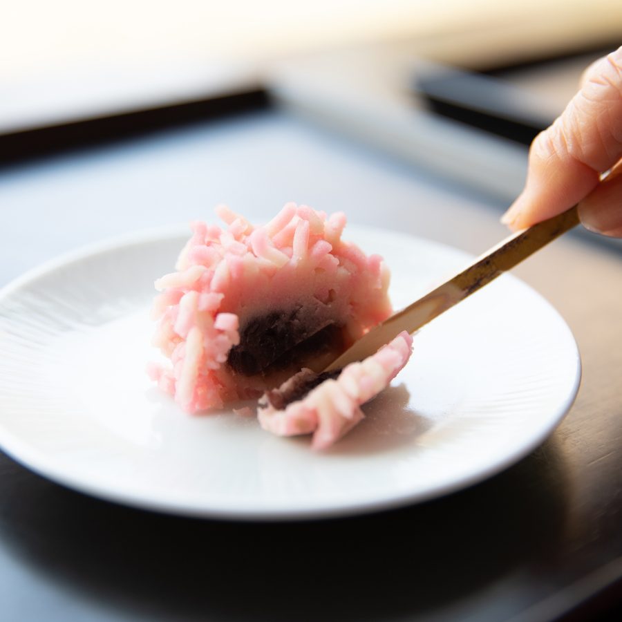 A closeup of a hand holding a golden dessert scoop slicing through a piece of pink sakuramochi, a Japanese sweet confectionery found in Kyoto.