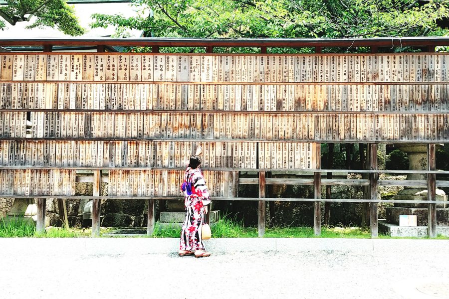 A woman in full kimono outfit touching wooden prayer boards in Kyoto on a sunny day.