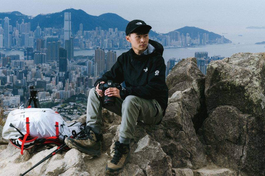 Hong Kong photographer Kelvin Yuen sitting on a rock, holding a camera, with a view of Kowloon seen from a the distance on top of Lion Rock