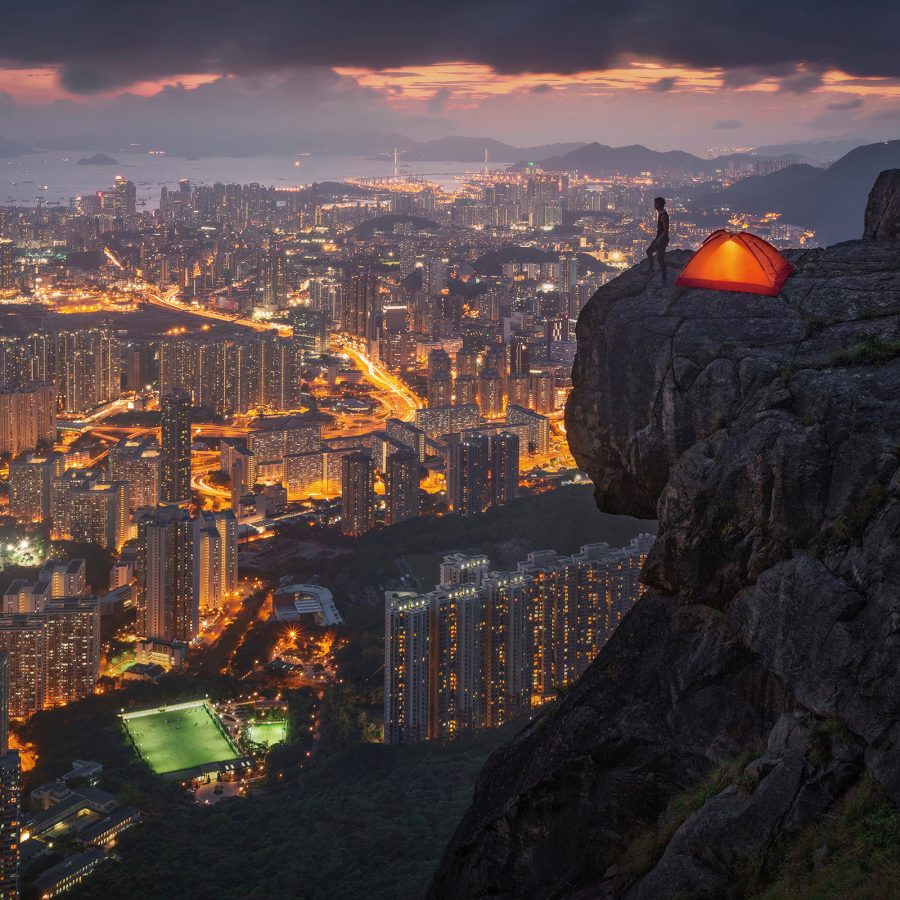 A drone shot of a man standing on the top of Lion Rock next to a camping tent overlooking the Kowloon skyline at night by Hong Kong photographer Kelvin Yuen