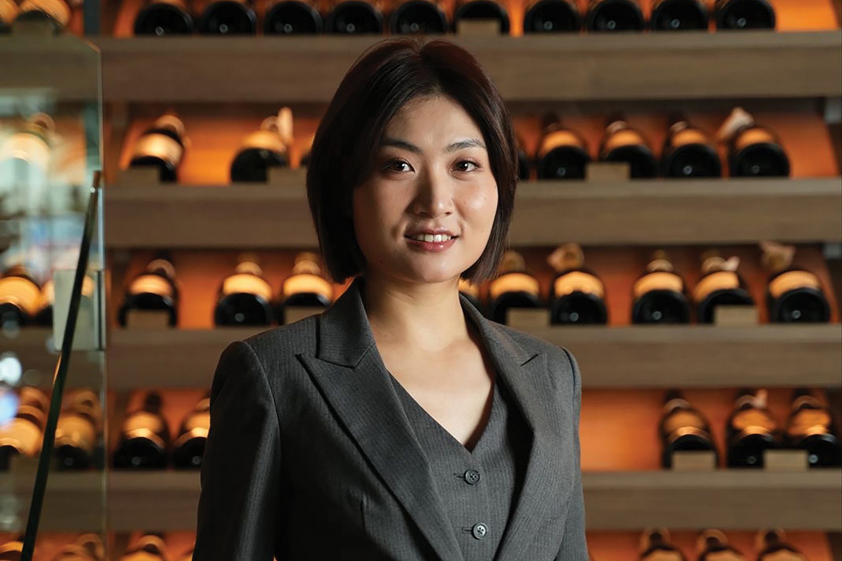 A portrait photo of wine expert and female sommelier Della Tang from Shenzhen posing in front of a wine cellar display full of wine bottles.