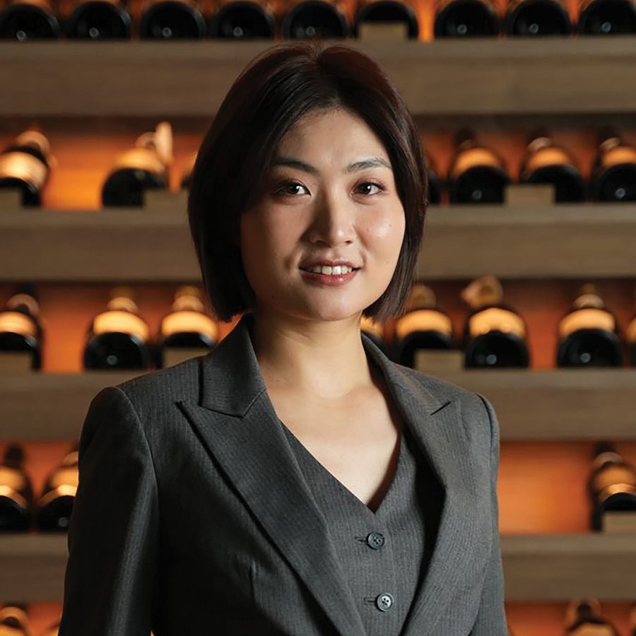 A portrait photo of wine expert and female sommelier Della Tang from Shenzhen posing in front of a wine cellar display full of wine bottles.