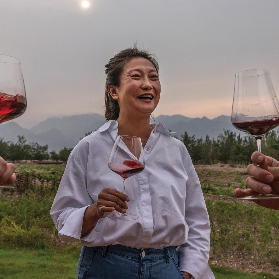 Female Chinese winemaker and founder of Silver Heights Vineyard laughing while holding a glass of red wine outdoors at a vineyard on a sunny day with two hands holding up glasses of red wine from each side.
