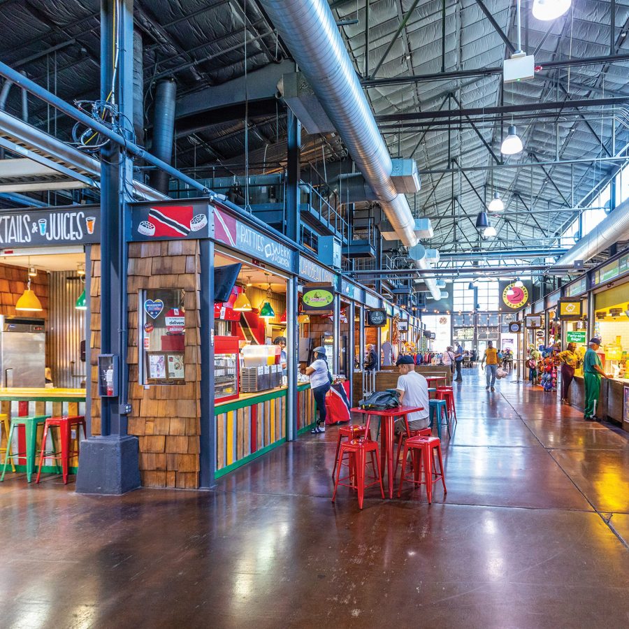 The interior of Dallas Farmers Market includes dozens of shopfronts along a wide aisle, while colourful chairs are placed in front of an eatery. 