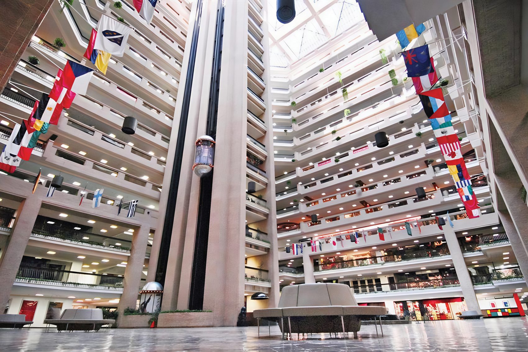 The interior of World Trade Center features an imposing and tall lobby surrounded by several floors of wholesale shopping, accessible by an outward facing elevator. National flags from around the world are also on display, extending outward from the first floor.