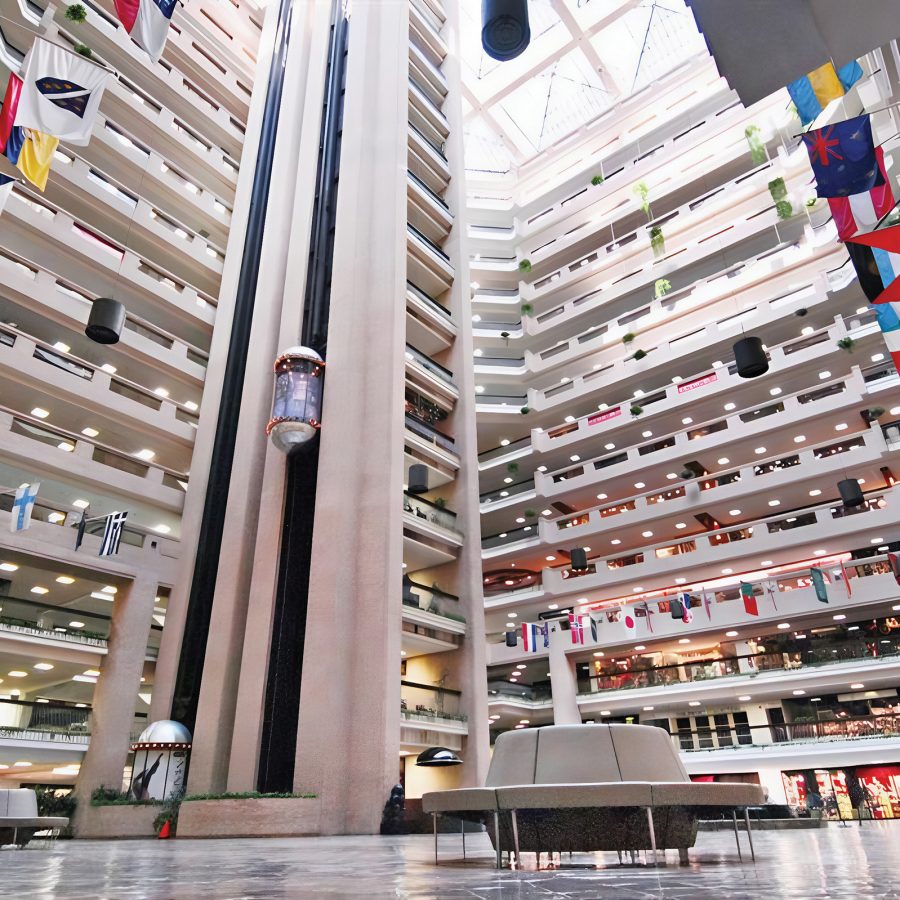 The interior of World Trade Center features an imposing and tall lobby surrounded by several floors of wholesale shopping, accessible by an outward facing elevator. National flags from around the world are also on display, extending outward from the first floor.
