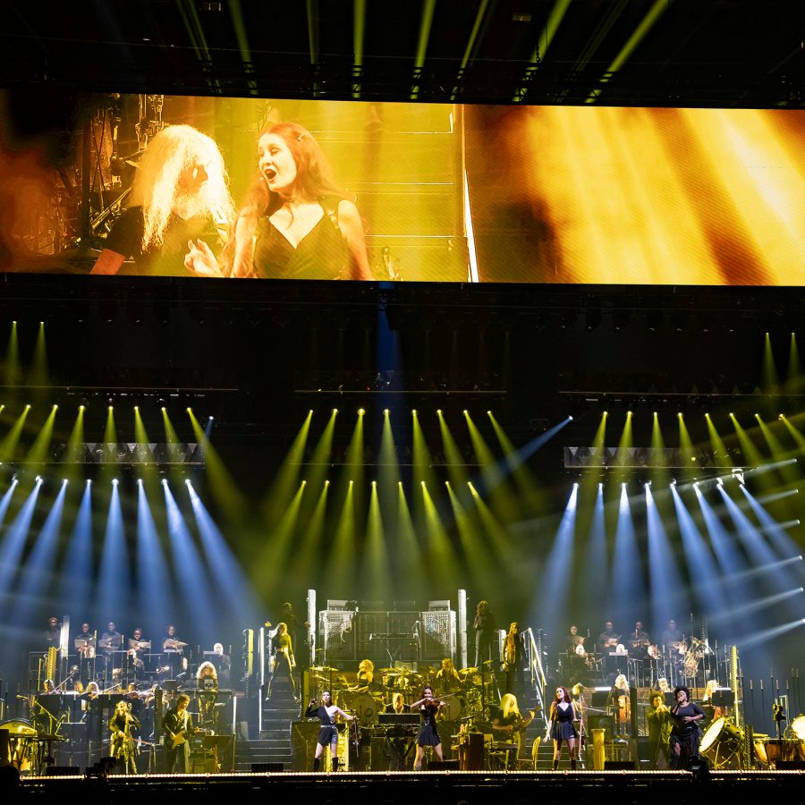 An orchestra on a stage is illuminated by spotlights as a screen above plays a scene from a movie scored by Hans Zimmer. 