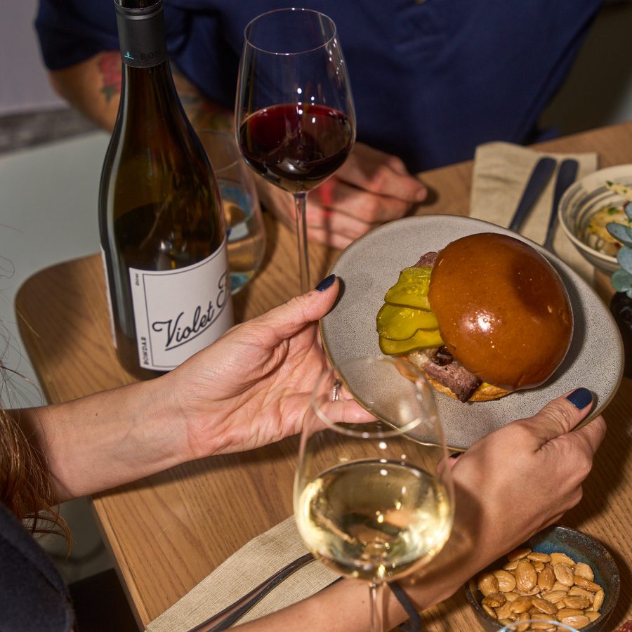 Two people dining with wine while a pair of hands hold up a plate of burger served at Servo in Hong Kong.