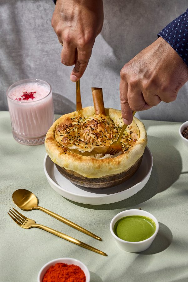 A vibrant shot of a person cutting into a lamb-shank biryani over a green table cloth with a pink cocktail in the background served at Spice Bazaar in Hong Kong.