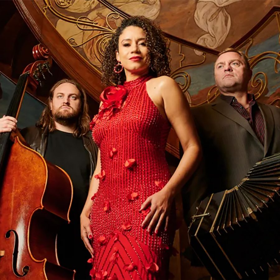   Singer Stephany Ortega stands by an ornate staircase, wearing a red tango dress. To her left a man stands with a large double bass; to her left a man stands with a large accordion.