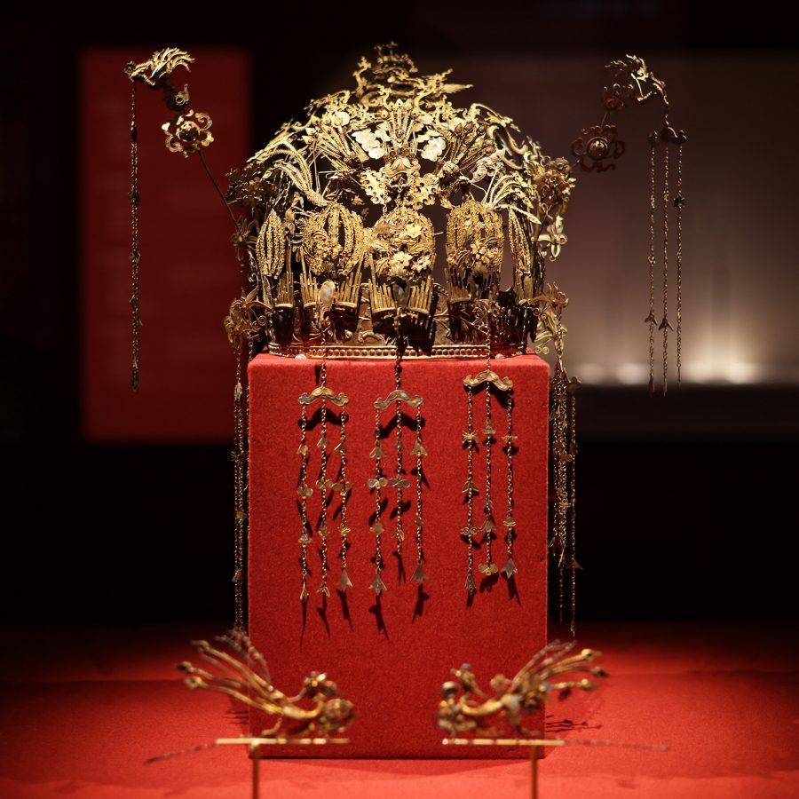 A large, elaborate piece of gold headwear is displayed on a red pedestal and lit by a spotlight at Guangdong Museum.