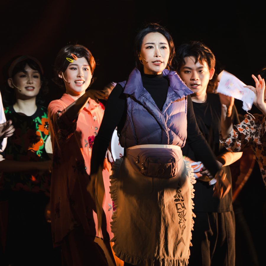 A scene from the stage drama I Am What I Am shows an ensable cast standing in a line against a black backdrop.